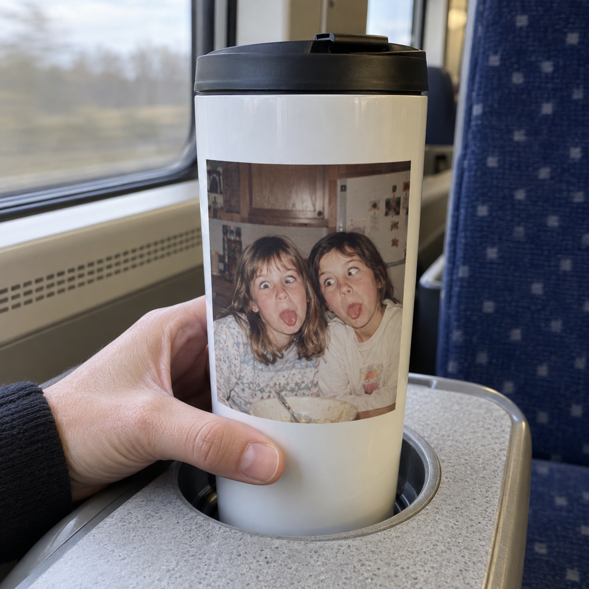 A white travel mug printed with a funny old photograph of sisters in a kitchen, shown on a train.