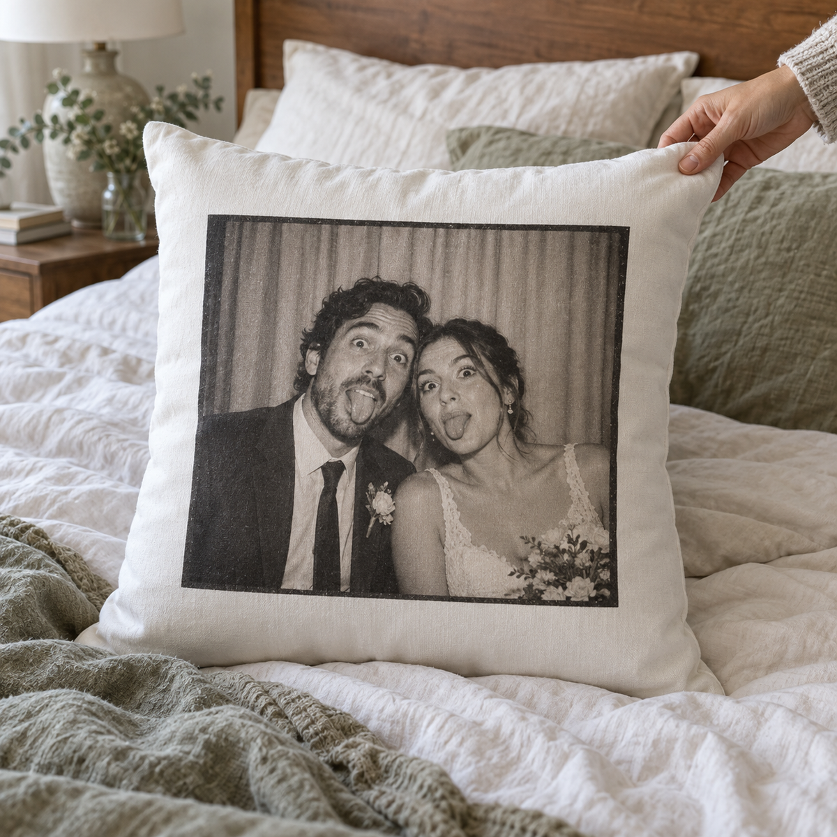 A white photo pillow printed with a funny wedding photo booth portrait, placed on an unmade bed.
