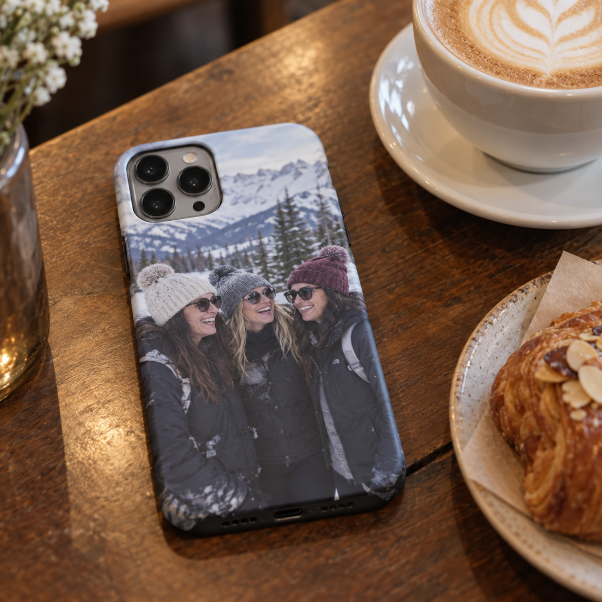 A slim personalized phone case printed with three friends on a past snowy trip, shown on a café table.