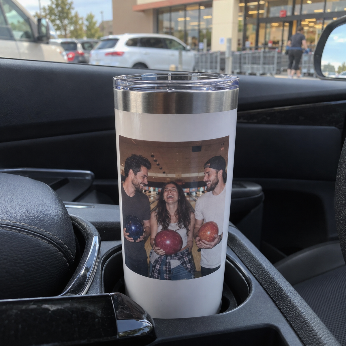 A stainless travel tumbler printed with a bowling photo of three friends, resting in a parked car cupholder.