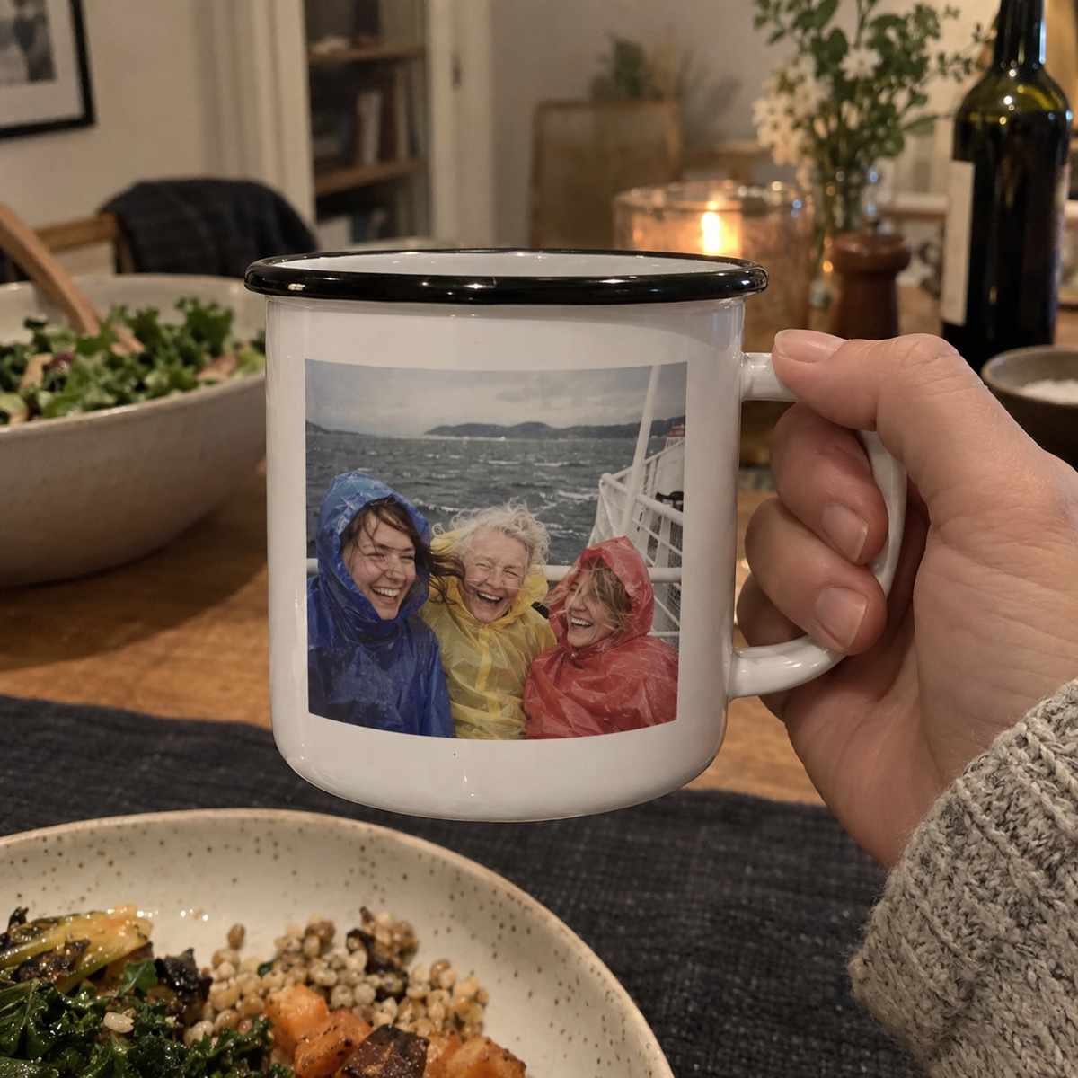Enamel camping mug printed with a windy ferry trip photo, held at an indoor dinner table.